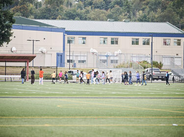 Group of many kids playing on a school turf soccer field with the school in the background.